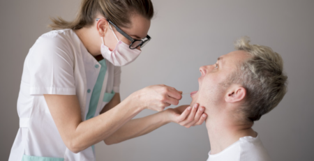 Nurse wearing a mask and glasses swabs a man's throat for a medical test in a clinical setting.
