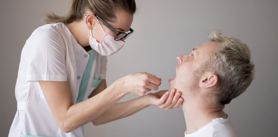 Nurse wearing a mask and glasses swabs a man's throat for a medical test in a clinical setting.