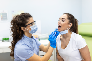 Dentist in a mask and face shield, wearing blue gloves, examines a patient’s mouth during a dental procedure.