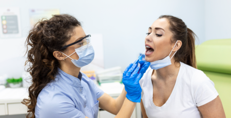 Dentist in a mask and face shield, wearing blue gloves, examines a patient’s mouth during a dental procedure.
