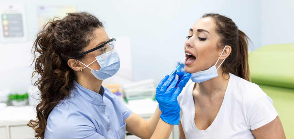 Dentist in a mask and face shield, wearing blue gloves, examines a patient’s mouth during a dental procedure.