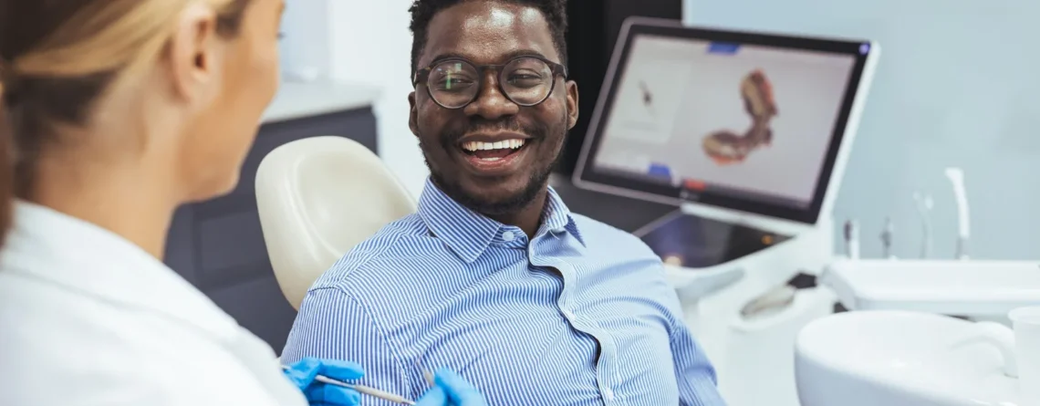 Smiling man in a blue striped shirt sits in a dental chair while a dentist in gloves prepares a syringe.