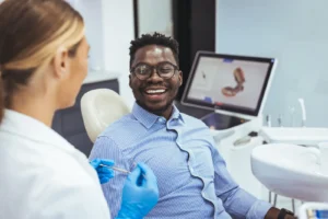 Smiling man in a blue striped shirt sits in a dental chair while a dentist in gloves prepares a syringe.