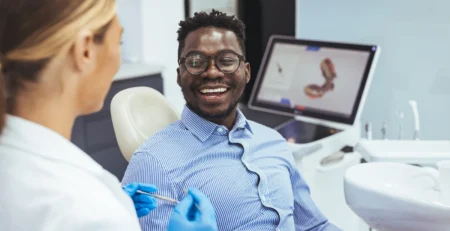 Smiling man in a blue striped shirt sits in a dental chair while a dentist in gloves prepares a syringe.