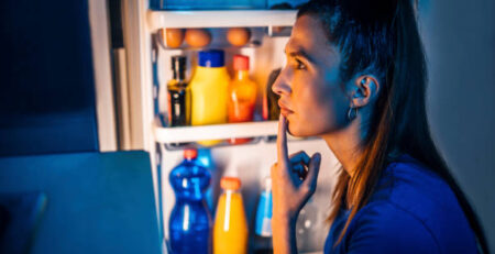 Woman standing at an open refrigerator at night deciding whether eating before bed is bad
