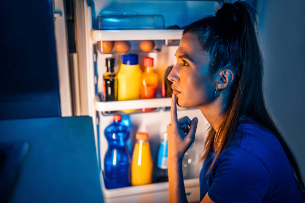 Woman standing at an open refrigerator at night deciding whether eating before bed is bad